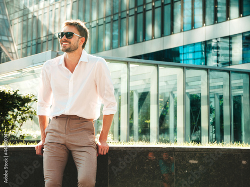 Portrait of handsome smiling stylish hipster lambersexual model.Modern man dressed in white shirt. Fashion male posing in the street background near skyscrapers in sunglasses