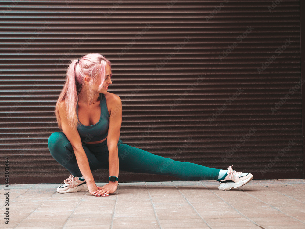 Obraz premium Fitness smiling woman in green sports clothing with pink hair. Young beautiful model with perfect body.Female posing in the street near roller shutter wall. Stretching out before training