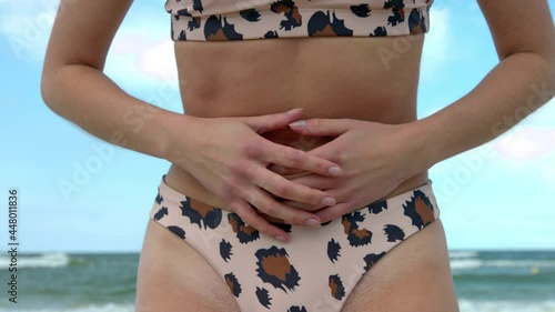 A young adult lady in a bikini standing at the beach got a rough tummy pain. A woman in a bikini with a stomachache bending her back and her hands pressing the tummy. A woman with a slim painful tummy photo