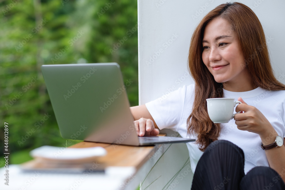 A beautiful young asian woman drinking coffee while using and working on laptop computer in the outdoors A beautiful young asian woman drinking coffee while using and working on laptop computer in the outdoors