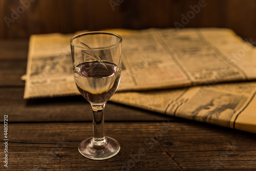Glass of vodka and newspaper on an old wooden table. Angle view, focus on the glass of vodka