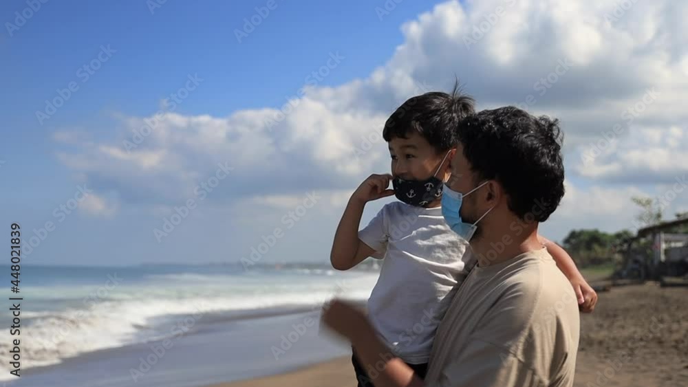 Close up of loving father straighten medical mask on his son face while holding him. Happy family enjoy a beautiful view of summer beach Close up of loving father straighten medical mask on his son face while holding him. Happy family enjoy a beautiful view of summer beach