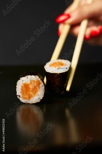 One sushi roll rests on a mirrored black surface and the other is raised by sushi sticks. You can see a woman's hand with a manicure.