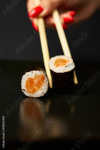 One sushi roll rests on a mirrored black surface and the other is raised by sushi sticks. You can see a woman's hand with a manicure.