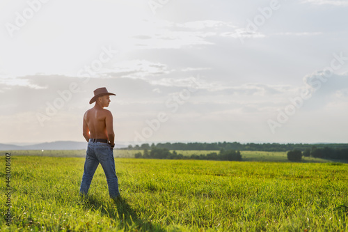 A man in a cowboy hat and jeans stands in a field beyond the sunset. Evening landscape in warm colors on the farm.