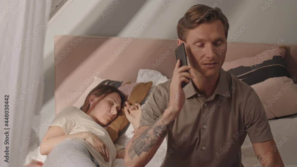Waist-up of young Caucasian man sitting on bed at home, talking on cellphone quietly, checking on his girlfriend sleeping on background Waist-up of young Caucasian man sitting on bed at home, talking on cellphone quietly, checking on his girlfriend sleeping on background