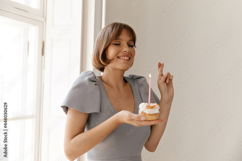 Birthday girl making wish at light room. Cute dark-haired lady with pale skin and nude makeup, wearing blue dress, smiling, crossing fingers and holding piece of cake Birthday girl making wish at light room. Cute dark-haired lady with pale skin and nude makeup, wearing blue dress, smiling, crossing fingers and holding piece of cake