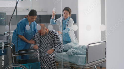 Canvas Print Medical team helping sick patient with leg injury fracture putting in wheelchair going on physiotherapy recovery in hospital ward