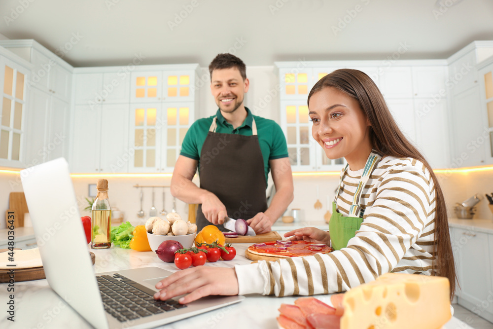 Happy couple making pizza together while watching online cooking course via laptop in kitchen Happy couple making pizza together while watching online cooking course via laptop in kitchen