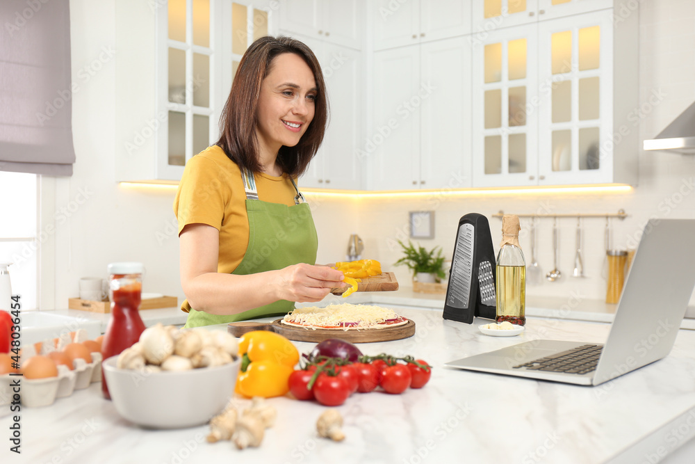 Woman making pizza while watching online cooking course via laptop in kitchen