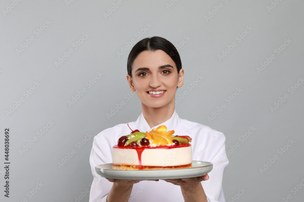 Happy professional confectioner in uniform holding delicious cake on light grey background Happy professional confectioner in uniform holding delicious cake on light grey background