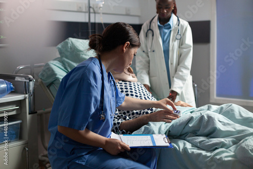 Картината върху платно Medical assistant checking oximeter attached to senior man laying in hospital bed, monitoring pacient and african doctor discussing with sick hospitalized senior man
