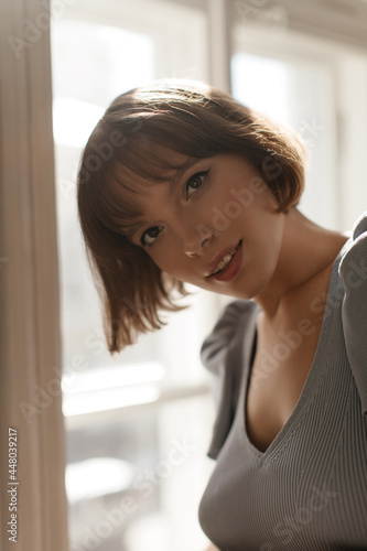 Lifestyle portrait of young curly girl looking into camera. Elegant short-haired woman with nude makeup and light blue top, posing at light room against window