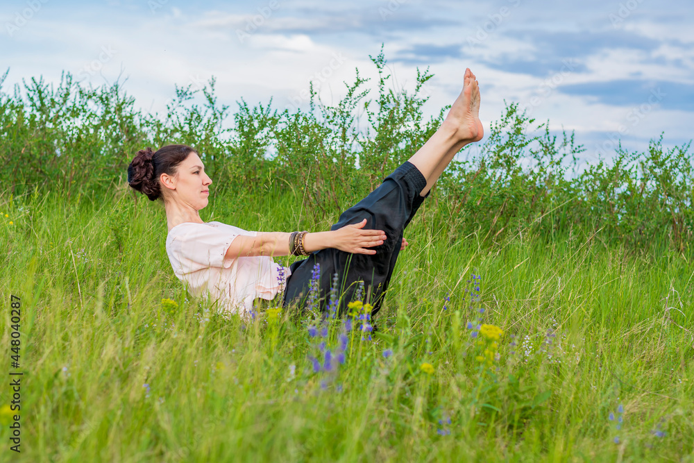 Beautiful young woman practices yoga outdoors. Sits on the ground with legs raised and holds a corner. Beautiful young woman practices yoga outdoors. Sits on the ground with legs raised and holds a corner.