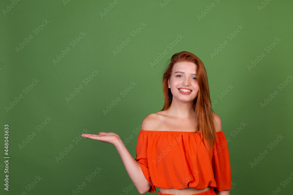Young red hair woman in casual orange blouse on green background smile and laugh holds empty space on hand Young red hair woman in casual orange blouse on green background smile and laugh holds empty space on hand