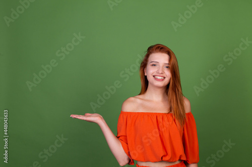 Young red hair woman in casual orange blouse on green background smile and laugh holds empty space on hand