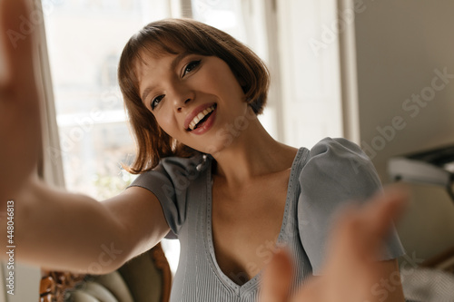 Good-quality selfie-portrait of cute girl at light room. Pretty young short-haired lady with nude makeup, toothy smile and blue dress, making photo and looking into camera Free Download Good-quality selfie-portrait of cute girl at light room. Pretty young short-haired lady with nude makeup, toothy smile and blue dress, making photo and looking into camera - GFXLoad