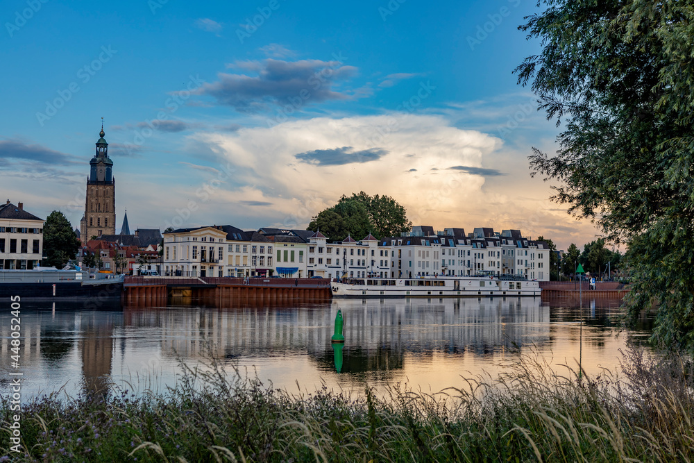 Countenance cityscape of Zutphen in The Netherlands with large cumulonimbus rain cloud reflecting in river IJssel seen from the opposite side framed by greenery. Dutch weather condition landscape.