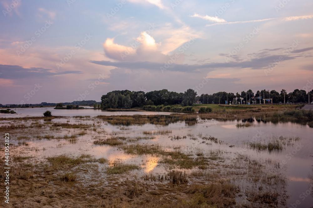 Floodplains and floodland half under water in Zutphen, The Netherlands, reflecting cumulus rain clouds above in colorful sunset tones. Dutch climate weather condition landscape concept.