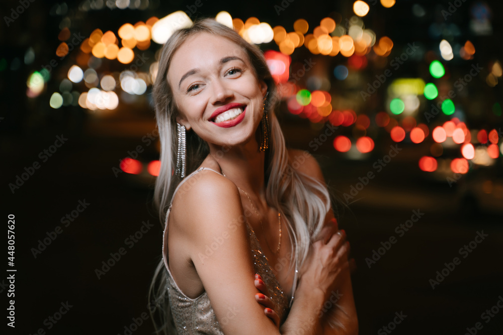 satisfied blonde woman smiling looking at camera at summer evening street. Stylish fancy golden cocktail Glided dress and long earrings. Summer party mood. Bokeh lights background. Red lips satisfied blonde woman smiling looking at camera at summer evening street. Stylish fancy golden cocktail Glided dress and long earrings. Summer party mood. Bokeh lights background. Red lips
