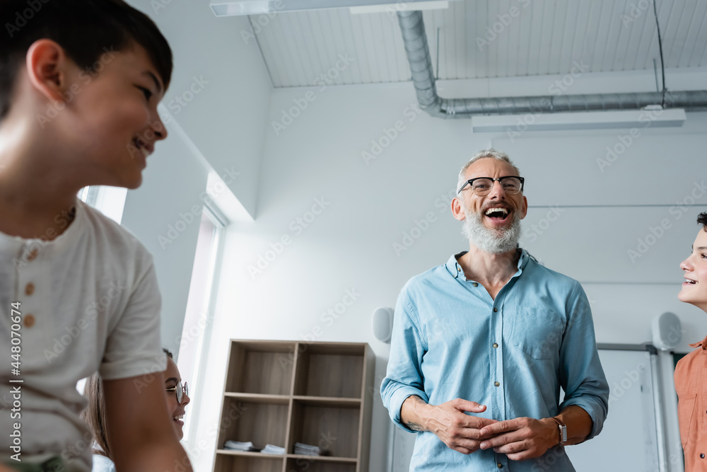 low angle view of bearded teacher laughing near multiethnic pupils in classroom low angle view of bearded teacher laughing near multiethnic pupils in classroom