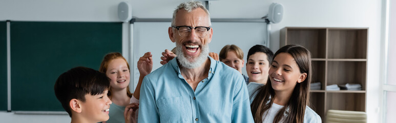 excited middle aged teacher laughing with closed eyes near happy multicultural pupils, banner