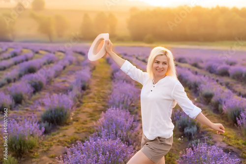 Fototapeta Naklejka Na Ścianę i Meble -  Beautiful young healthy woman with a white dress running joyfully through a lavender field holding a straw hat under the rays of the setting sun