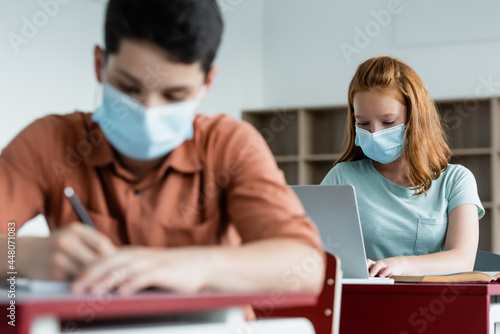 Schoolkid in medical mask using laptop near blurred classmate