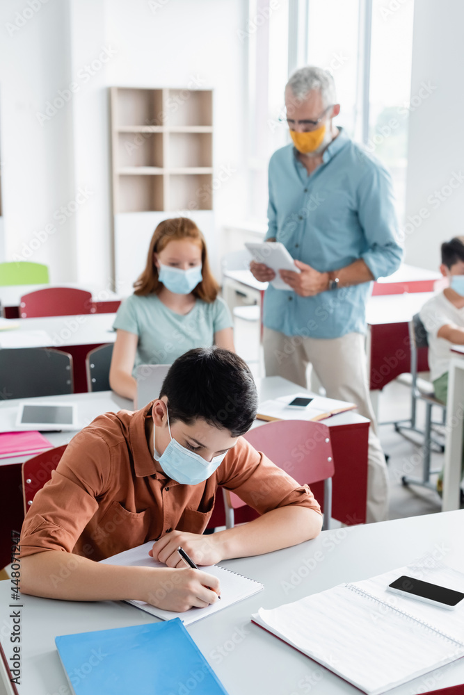 Schoolkid in medical mask writing on notebook in classroom Schoolkid in medical mask writing on notebook in classroom