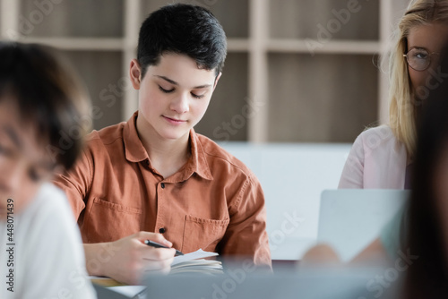 Schoolboy holding pen near notebook and blurred multiethnic pupils