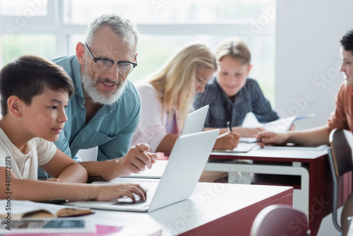 Mature teacher pointing at laptop near asian pupil in classroom