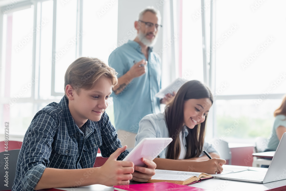 Smiling schoolboy using digital tablet near girl writing on notebook and blurred teacher Smiling schoolboy using digital tablet near girl writing on notebook and blurred teacher