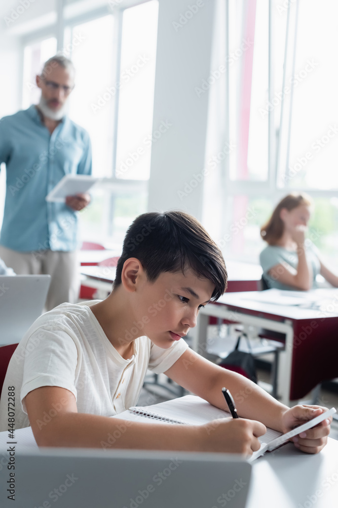 Fototapeta premium Asian schoolboy holding smartphone and writing on notebook in classroom