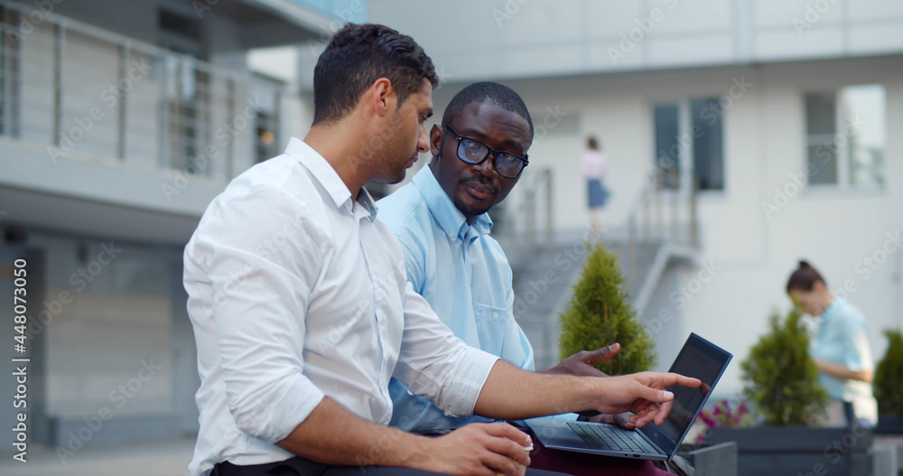 Multiethnic office colleagues using laptop sitting on bench outdoor. Multiethnic office colleagues using laptop sitting on bench outdoor.