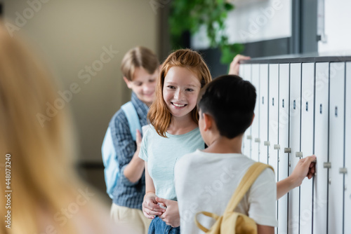 selective focus of redhead schoolgirl smiling in school corridor near blurred teenagers