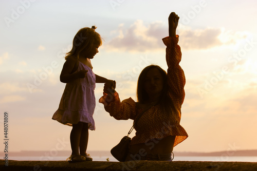 A silhouette of a happy mother and child against a yellow-orange sky at sunset. A dancing woman is holding a 2-3 year old baby girl, little kid by the hand. Maternal care, support, nurturing the baby.