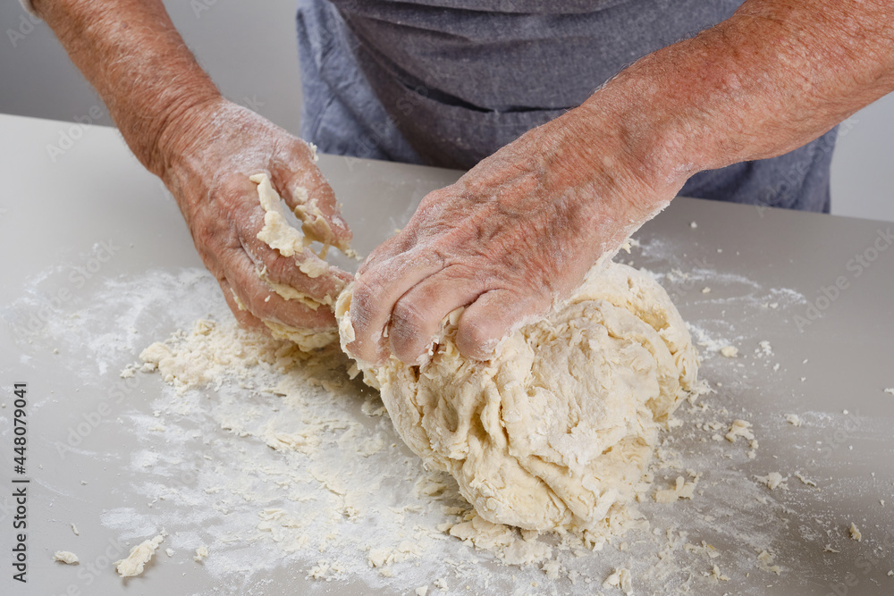 Female hands of an elderly woman knead the dough on a gray table.  Senior woman in a gray apron preparation of dough for a pizza, bread, pasta, festive cake. Selective focus