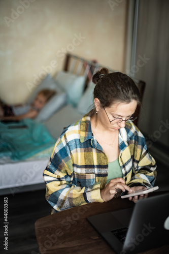 Front view of caucasian woman working on laptop from home, in cozy atmosphere, with flowers on the desk. Female wearing round eyeglasses is using smartphone. Her little daughter lie in bed behind