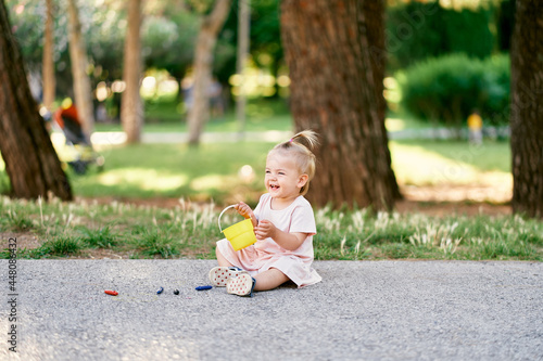 Little girl sitting on an asphalt path in a green park with a toy bucket in her hands