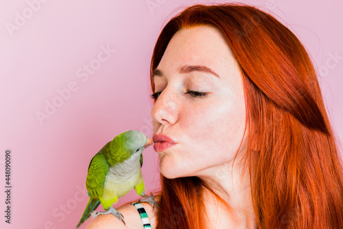A less-haired girl kisses a green monk parrot who sits on her shoulder on a pink background. photo