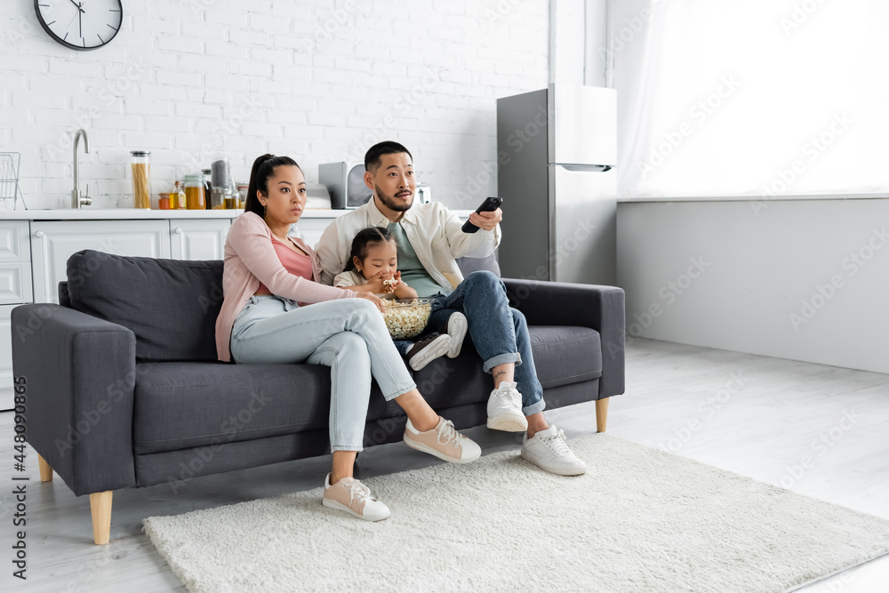focused asian family watching movie and kid eating popcorn in living room focused asian family watching movie and kid eating popcorn in living room