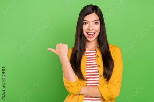 Photo of shiny excited young lady wear yellow shirt smiling pointing thumb empty space isolated green color background