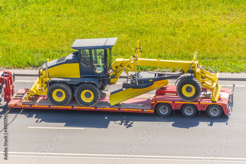 Grader tractor on transportation trailer truck with long trailer platform on the highway in the city.