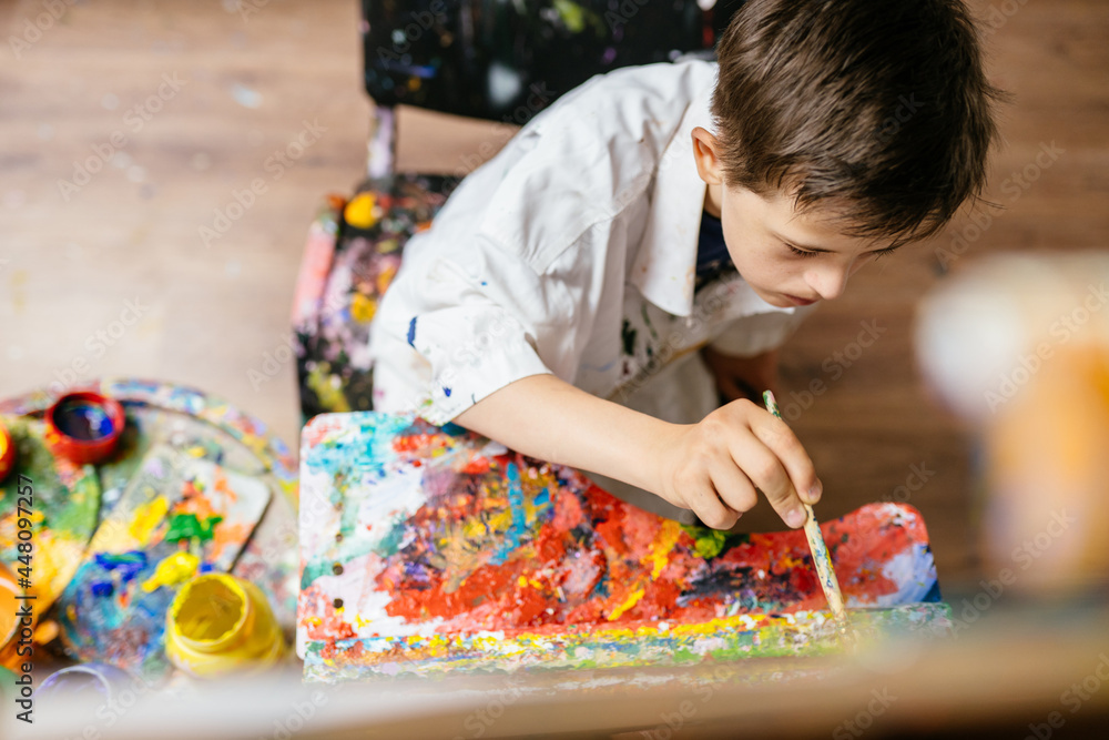 Young Caucasian down syndrome boy sitting at chair with colors and painting on easel with concentrated and calm painting. Concept for education for disabled kid. Young Caucasian down syndrome boy sitting at chair with colors and painting on easel with concentrated and calm painting. Concept for education for disabled kid.