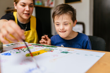 Young female teacher explain, talk at desk with a down syndrome schoolboy. Color painting for disabled kids, child who are down syndrome and student teacher, development of children with spesial needs