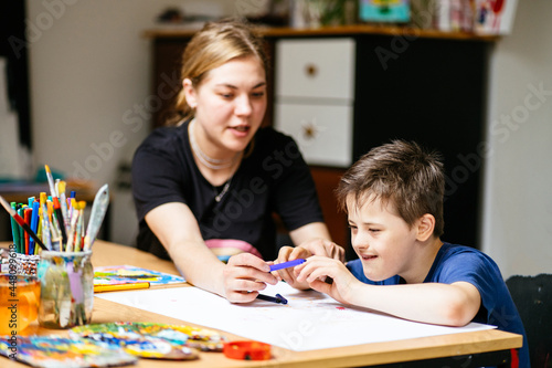 Close up of young female teacher sitting at desk with a Down syndrome schoolboy. Color painting on the paper for disabled kids, autism childs who are down syndrome and student teacher.