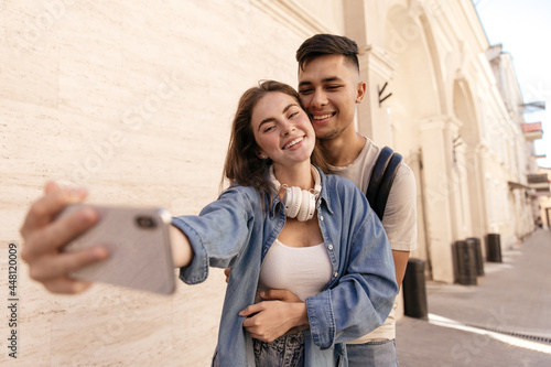 Joyful young couple making selfie-portrait in sunny city. Gorgeous dark-haired man smiling and hugging girlfriend, wearing white top and headphones, blue shirt