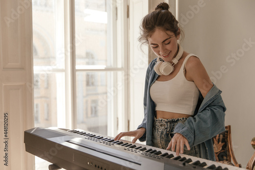 Cute young student playing piano at home near window. Attractive girl with collected hair, white top and blue shirt, headphones, practicing in music and smiling