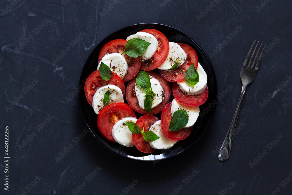 Italian caprese salad with fork on the black plate with copy space. sliced red tomatoes, mozzarella cheese, green basil, olive oil on a dark background. fresh salad.top view.