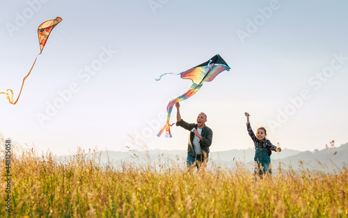 Outdoor photo of a father with daughter as they releasing colorful kites on the high grass meadow sincerely laughing. Warm family moments or outdoor time spending concept image.